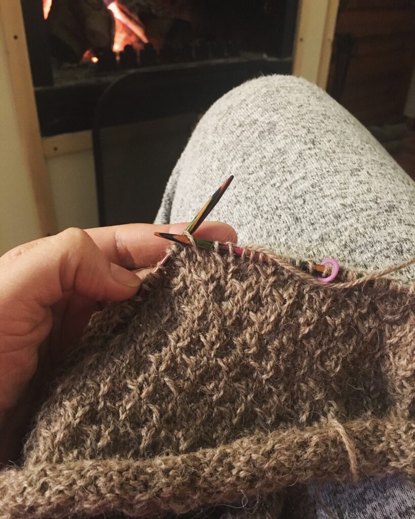 Proyecto de tejido a medio hacer, frente a una chimenea encendida, en un ambiente cálido y relajante ideal para momentos de autocuidado.